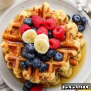 Protein-Packed Strawberry Banana Waffles 3 A stack of waffles topped with blueberries, raspberries, banana slices, and drizzled with syrup on a round white plate.