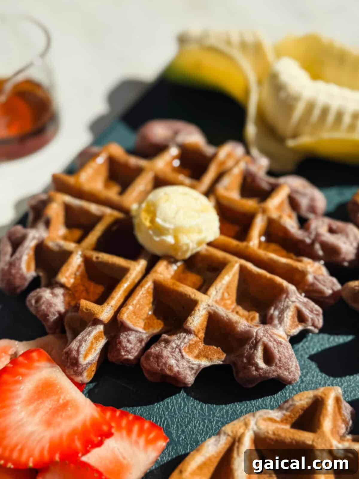 Protein-Packed Strawberry Banana Waffles 4 A close-up of a waffle topped with a scoop of butter and syrup, with sliced strawberries and a partially peeled banana in the background.