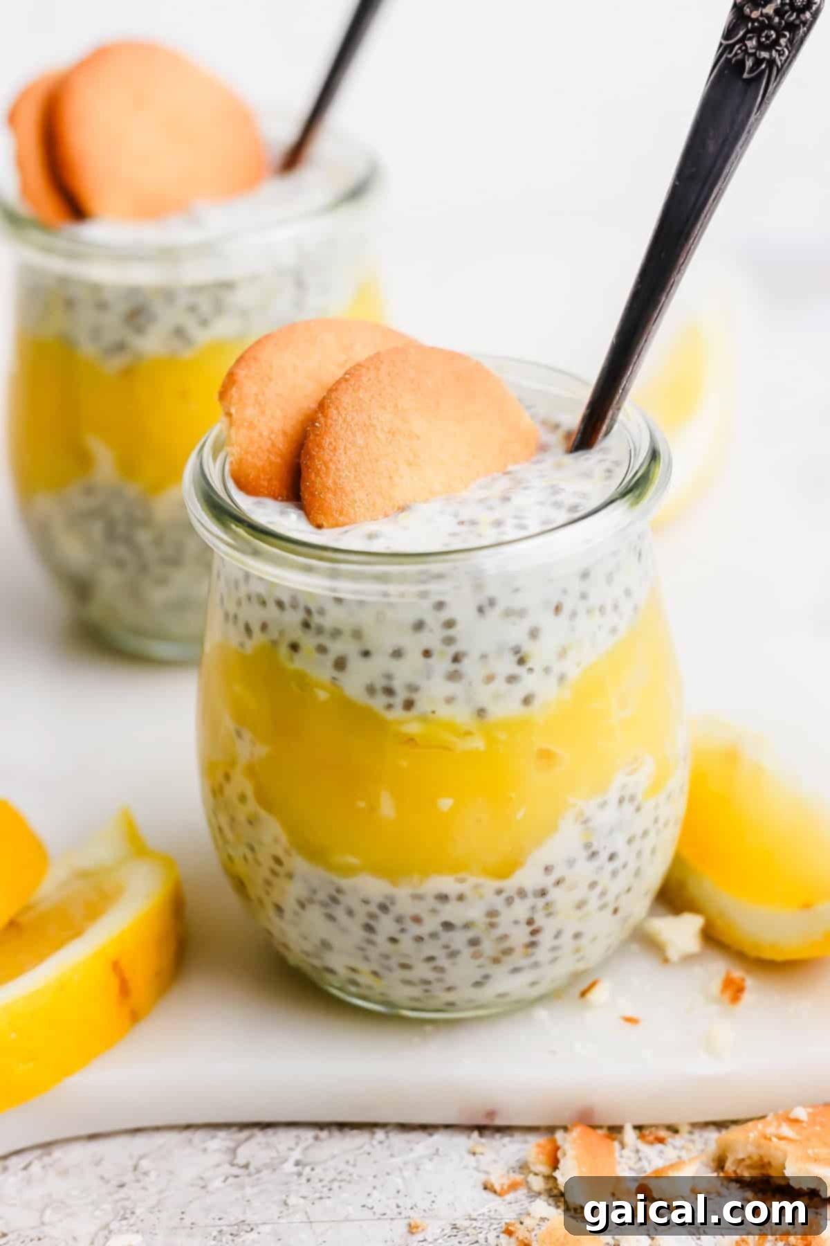 Protein-Packed Lemon Cream Pie Chia Pudding 2 Two jars of chia pudding layered with yellow lemon curd, topped with two cookies and a spoon, surrounded by lemon slices.