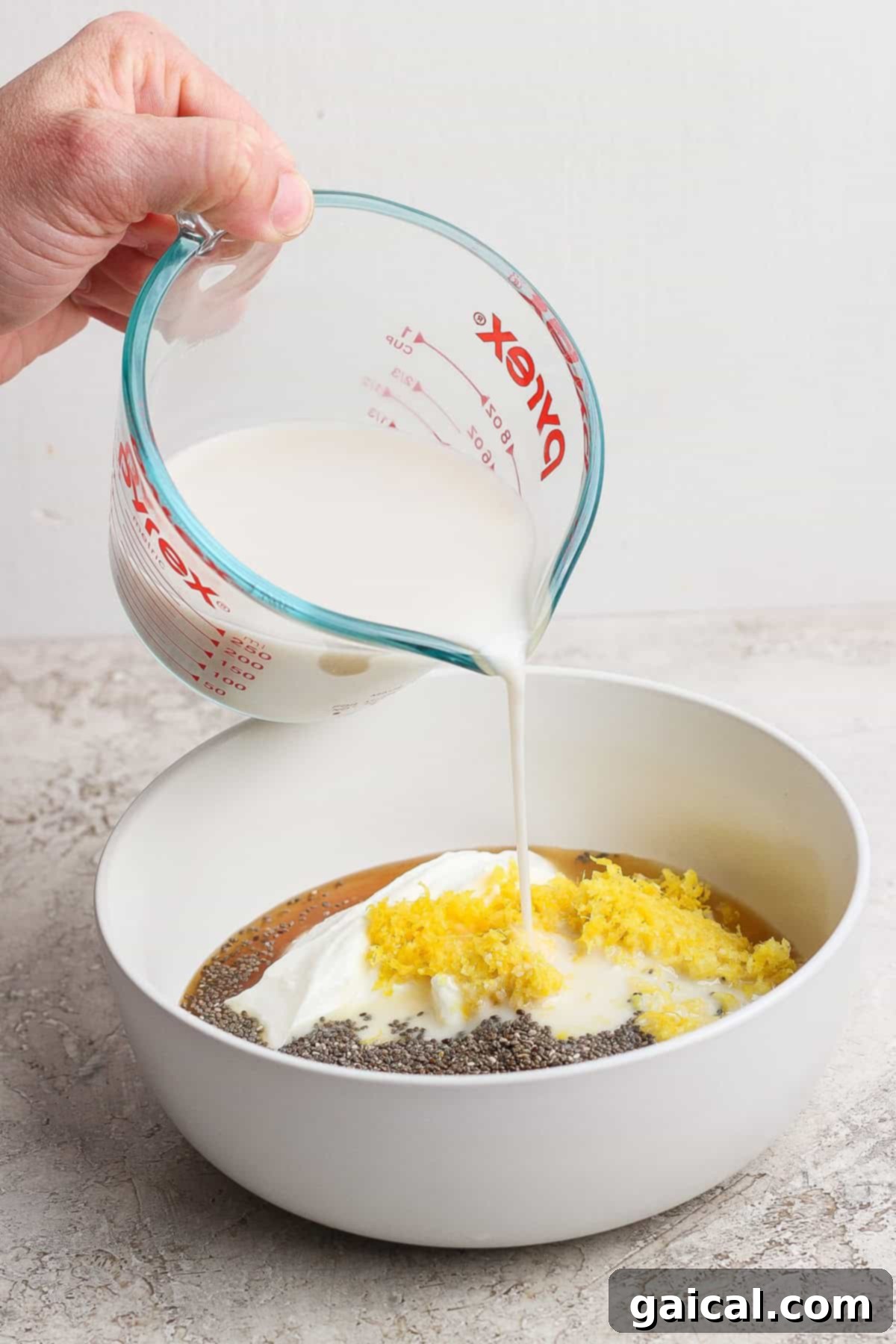 Protein-Packed Lemon Cream Pie Chia Pudding 3 Pouring milk from a measuring cup into a bowl containing yogurt, chia seeds, lemon zest, and liquid.