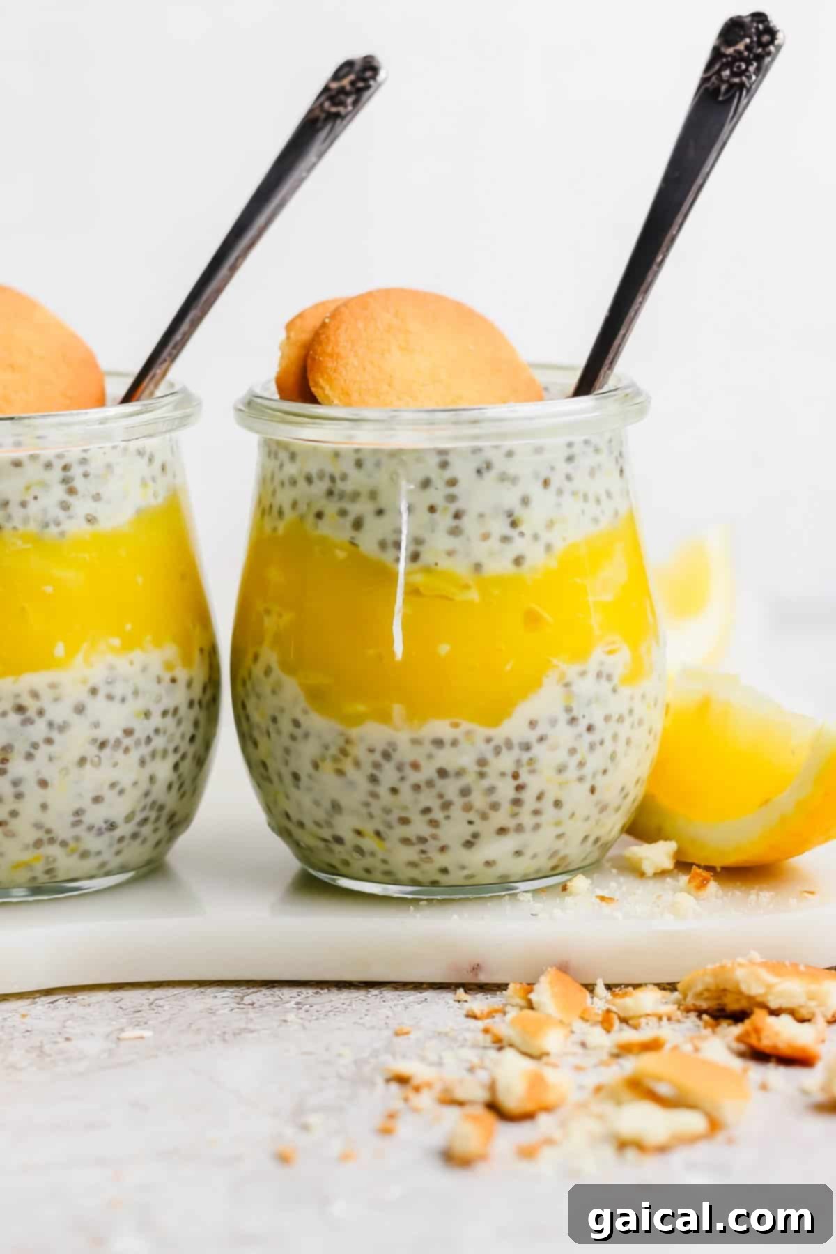 Protein-Packed Lemon Cream Pie Chia Pudding 4 Two jars of chia pudding layered with lemon curd, each garnished with a cookie, placed on a marble surface with lemon wedges and cookie crumbs nearby.