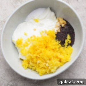 A bowl containing yogurt, lemon zest, poppy seeds, minced garlic, and salt on a light-colored surface.