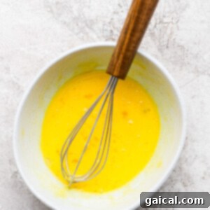 Whisk in a bowl of beaten eggs on a textured, light-colored surface.