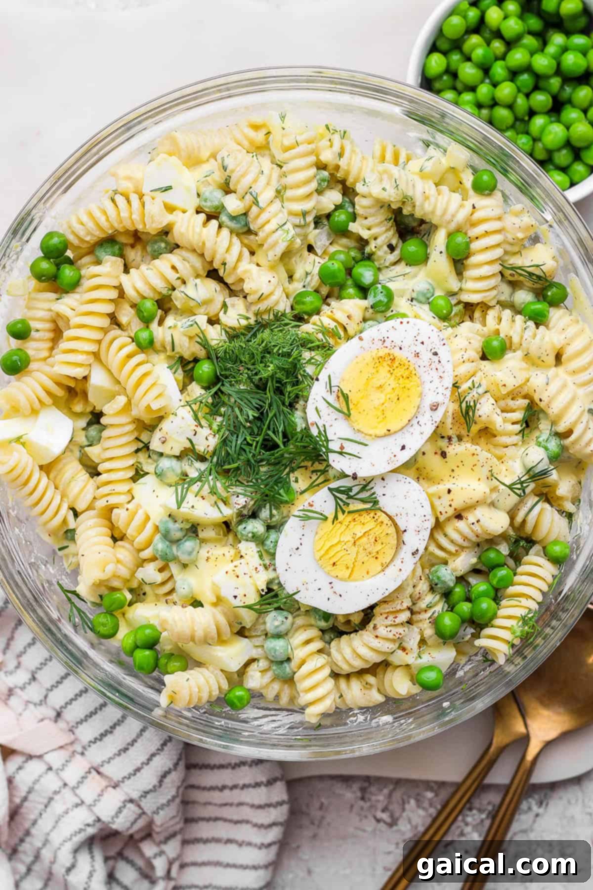 A vibrant bowl of creamy deviled egg pasta salad featuring rotini, bright green peas, slices of hard-boiled eggs, fresh dill, and a luscious dressing. A golden spoon and a neatly folded cloth napkin are placed beside the bowl, hinting at an inviting meal.