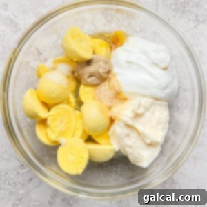 Bowl containing boiled egg yolks, mayonnaise, mustard, yogurt, and seasoning on a textured surface.