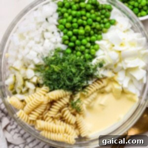 Glass bowl containing rotini pasta, peas, chopped onions, boiled eggs, pickles, fresh dill, and mayonnaise, set on a white surface with a silver spoon nearby.