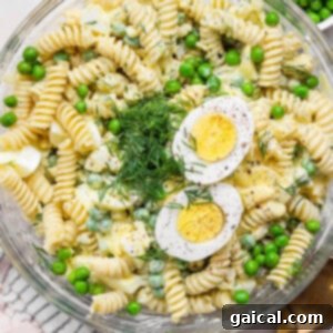 A bowl of pasta salad with rotini, green peas, hard-boiled egg slices, dill, and a creamy dressing. A gold spoon and a cloth napkin are next to the bowl.