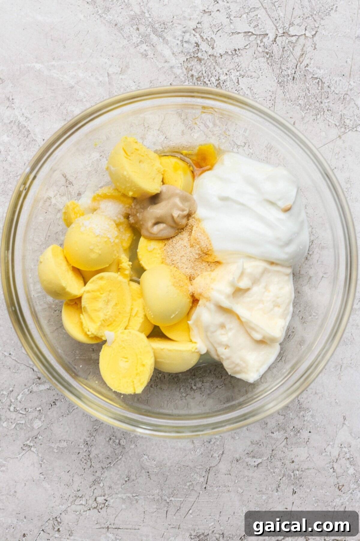 A glass bowl containing mashed boiled egg yolks, mayonnaise, Dijon mustard, Greek yogurt, and various seasonings, ready to be mixed into a creamy dressing. The textured surface provides a rustic backdrop.