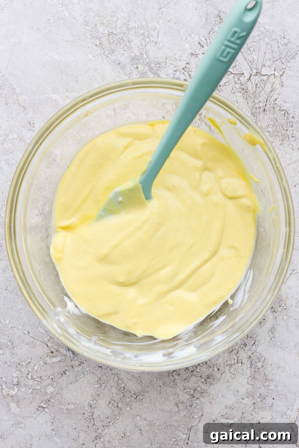 A glass bowl holds a perfectly smooth, creamy, and yellow deviled egg dressing, ready to be tossed with pasta and other ingredients. A teal spatula rests against the bowl on a textured gray surface.