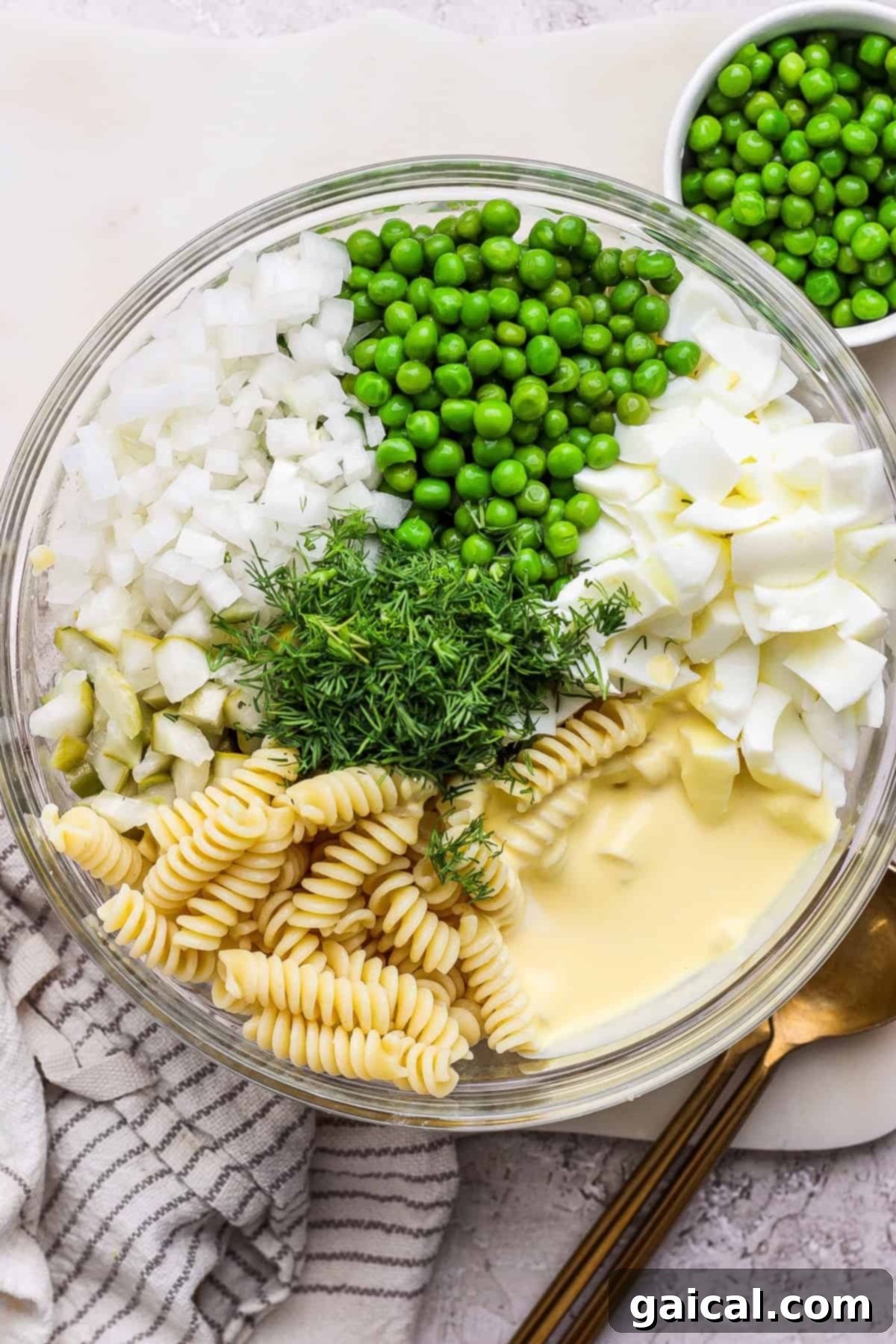 A spacious glass bowl brimming with a delightful deviled egg pasta salad, featuring plump rotini pasta, vibrant green peas, finely chopped onions, tender pieces of boiled egg, zesty pickles, and fresh dill, all generously coated in a creamy mayonnaise-based dressing. The bowl rests on a pristine white surface, with a glistening silver spoon casually placed nearby.