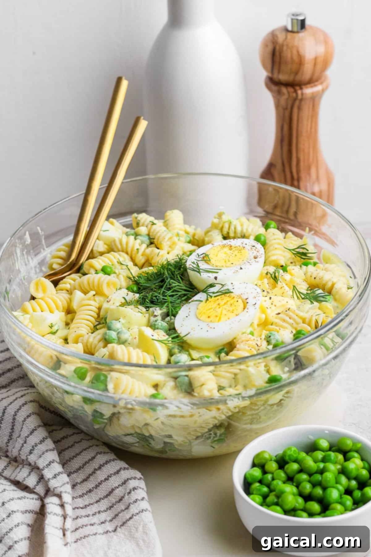 A beautifully arranged glass bowl of deviled egg pasta salad, featuring perfectly sliced hard-boiled eggs and a medley of fresh herbs, garnished with vibrant green peas. The bowl is artfully placed on a table alongside elegant chopsticks, a soft white towel, a small bowl of fresh peas, a sophisticated pepper mill, and a bottle of olive oil, suggesting a refined culinary experience.