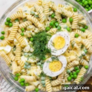 A bowl of fusilli pasta salad with green peas, chopped dill, and sliced hard-boiled egg halves on top.