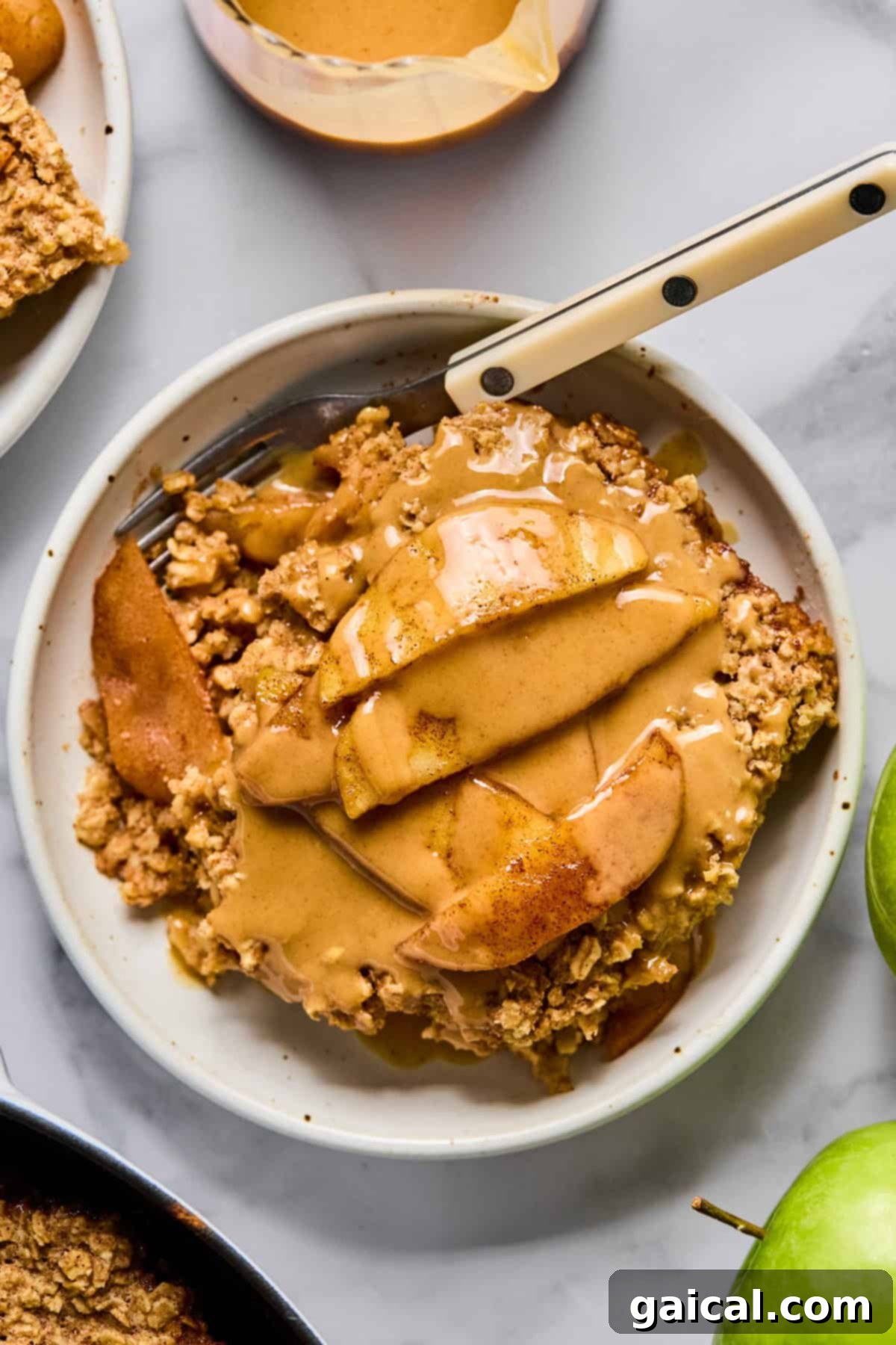 A slice of apple crumble topped with sliced apples and a drizzle of caramel sauce on a white plate with a fork, ready to be enjoyed.