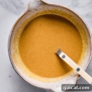 A ceramic bowl filled with a smooth, light brown liquid mixture and a white-handled spoon resting inside, placed on a marble surface, showing the making of the caramel sauce.