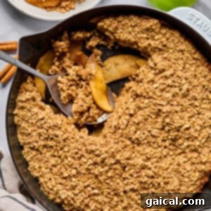 A cast iron skillet filled with baked apple crisp topped with oat crumble, with a serving scooped out onto a plate. Green apples and cinnamon sticks are nearby, completing the delicious presentation.