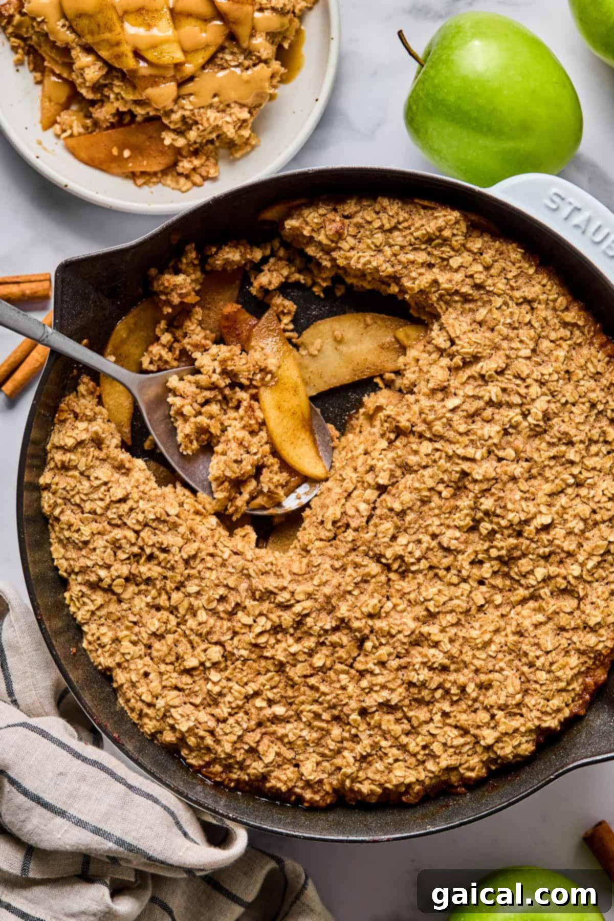 A cast iron skillet filled with baked apple crisp topped with oat crumble, with a serving scooped out onto a plate. Green apples and cinnamon sticks are nearby, creating an inviting scene.