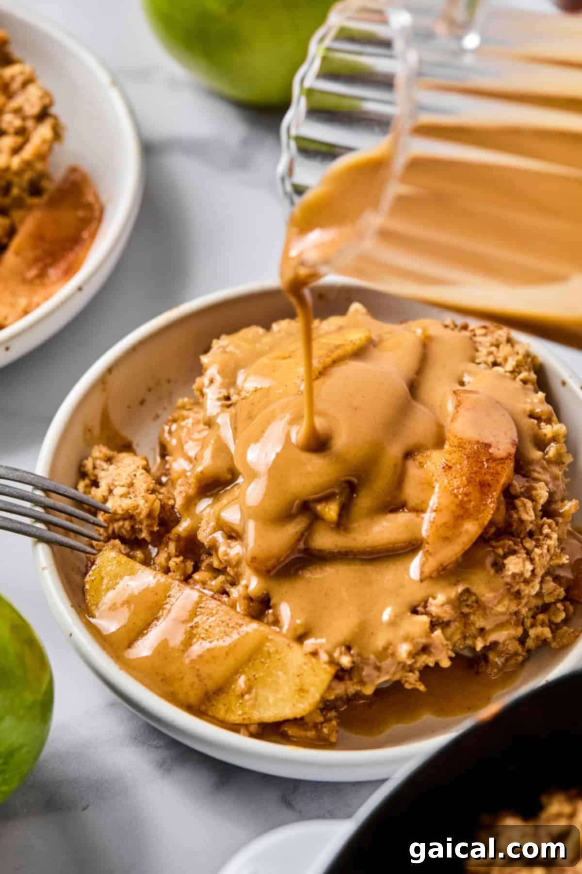 A bowl of apple crisp is topped with caramel sauce being poured from a glass container, with apples visible and a fork resting on the side, inviting a delicious taste.