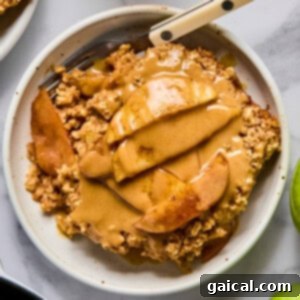 A bowl of apple crisp topped with sliced apples and a generous drizzle of nut butter, with a fork resting in the bowl.