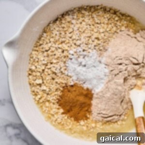 A mixing bowl containing oats, flour, baking powder, cinnamon, and a liquid ingredient with a spatula resting on the side, ready for mixing.