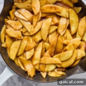 Sliced apples coated with cinnamon and sugar cooking in a cast iron skillet on a marble countertop, forming the base of the oatmeal bake.