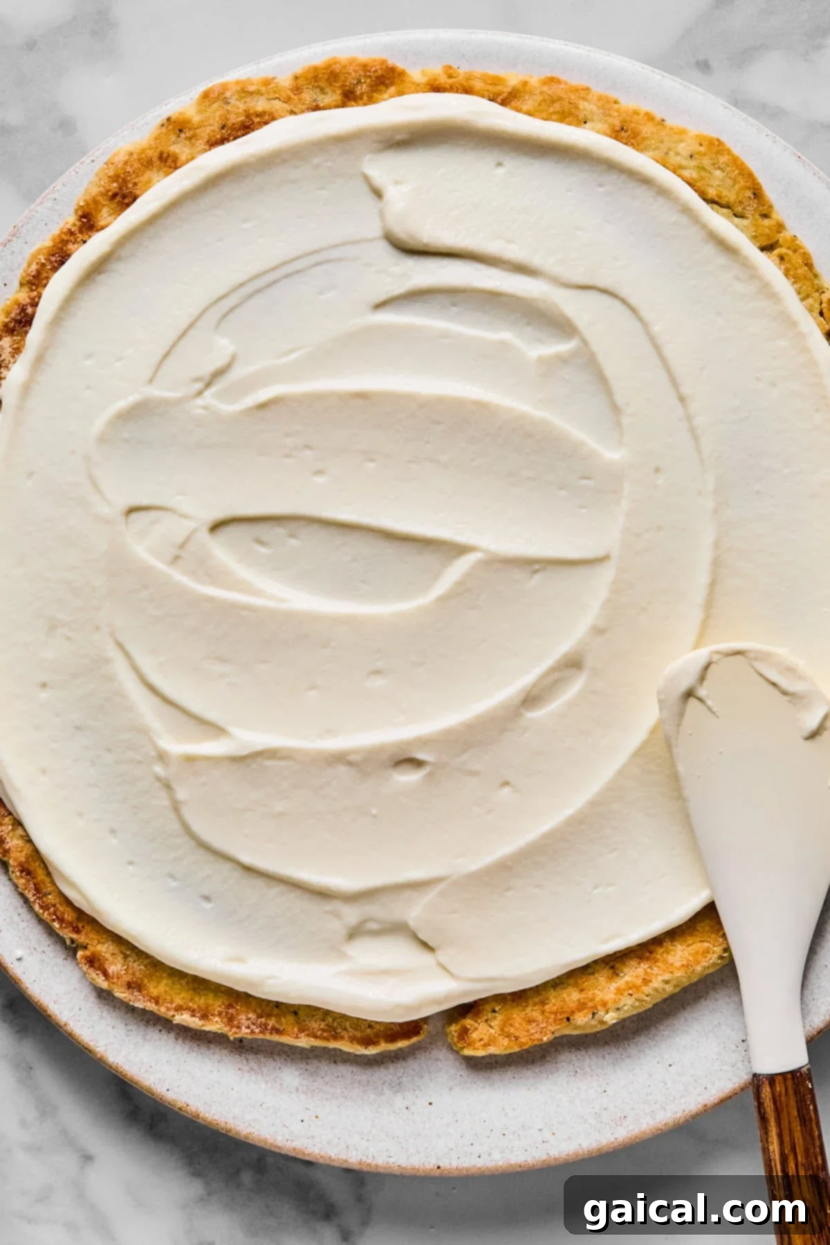 Protein-Packed Cottage Cheese Fruit Pizza 5 A smooth, creamy white frosting is being meticulously spread over a round cake on a white plate using an offset spatula, creating an even layer across the surface.