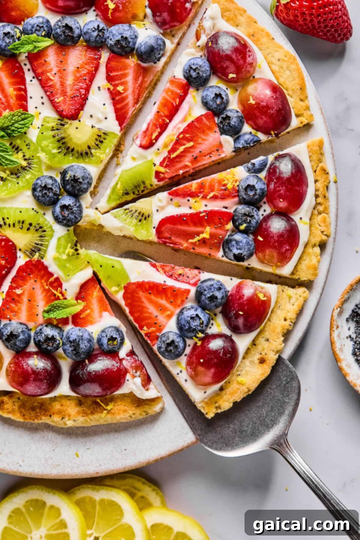 Protein-Packed Cottage Cheese Fruit Pizza 6 A beautifully arranged fruit pizza, garnished with fresh mint, showcasing a colorful assortment of sliced strawberries, red grapes, kiwi, and blueberries on a creamy white base. A pie server gently lifts one perfect slice, ready to be enjoyed.
