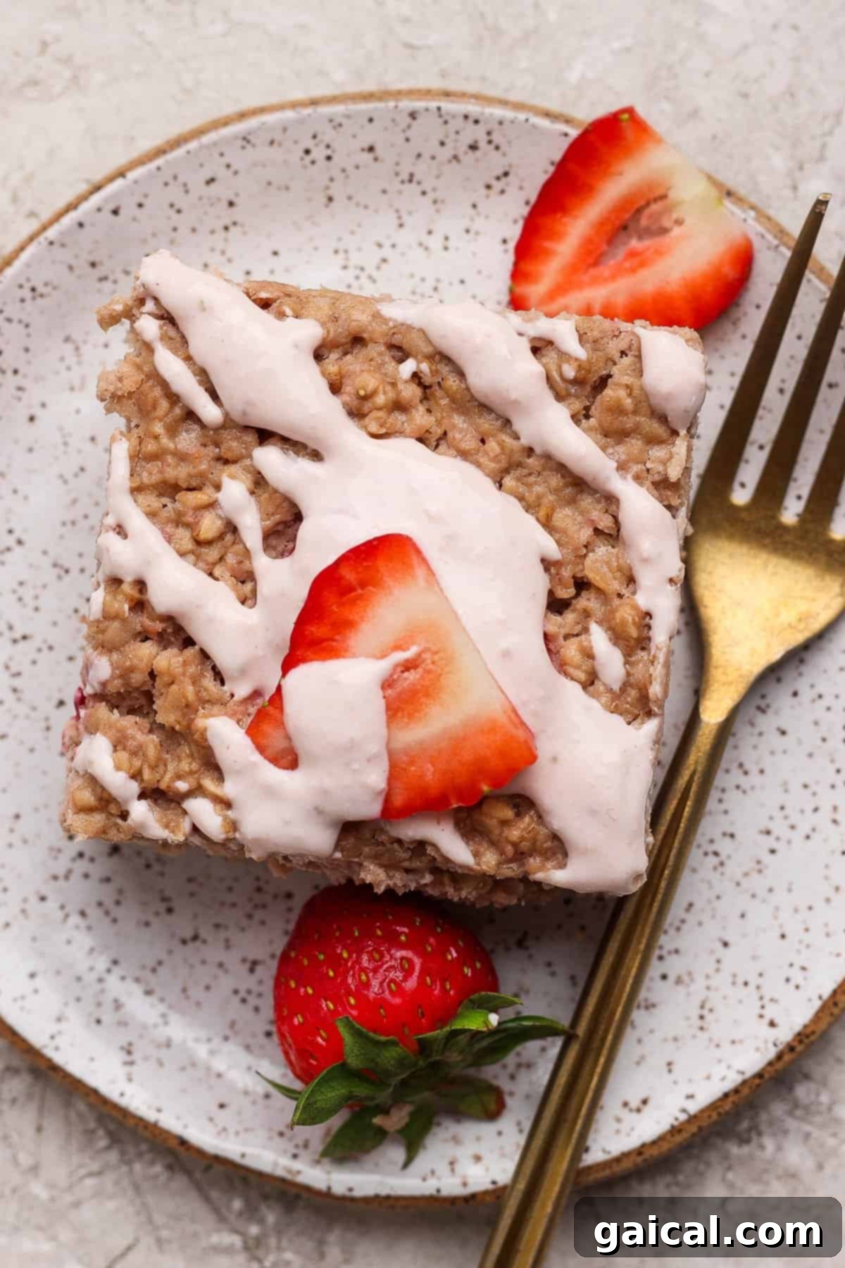 Creamy Strawberry Cheesecake Baked Oats 2 A square piece of strawberry cheesecake baked oatmeal topped with white icing and sliced fresh strawberries, served on a speckled plate with a gold fork beside it, inviting a delicious first bite.