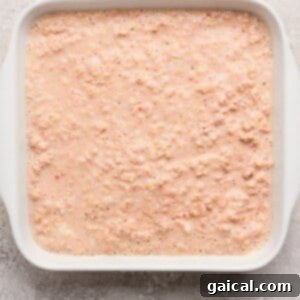 A white rectangular baking dish, meticulously filled with an uncooked, light pink oatmeal mixture, resting on a textured, light-colored surface, poised and ready for baking.