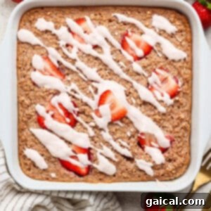 A square pan of baked oatmeal, beautifully adorned with vibrant sliced fresh strawberries and a creamy white icing drizzle, elegantly positioned on a light surface. Nearby, an arrangement of whole strawberries, a chic striped towel, and refined gold utensils create an inviting culinary tableau.