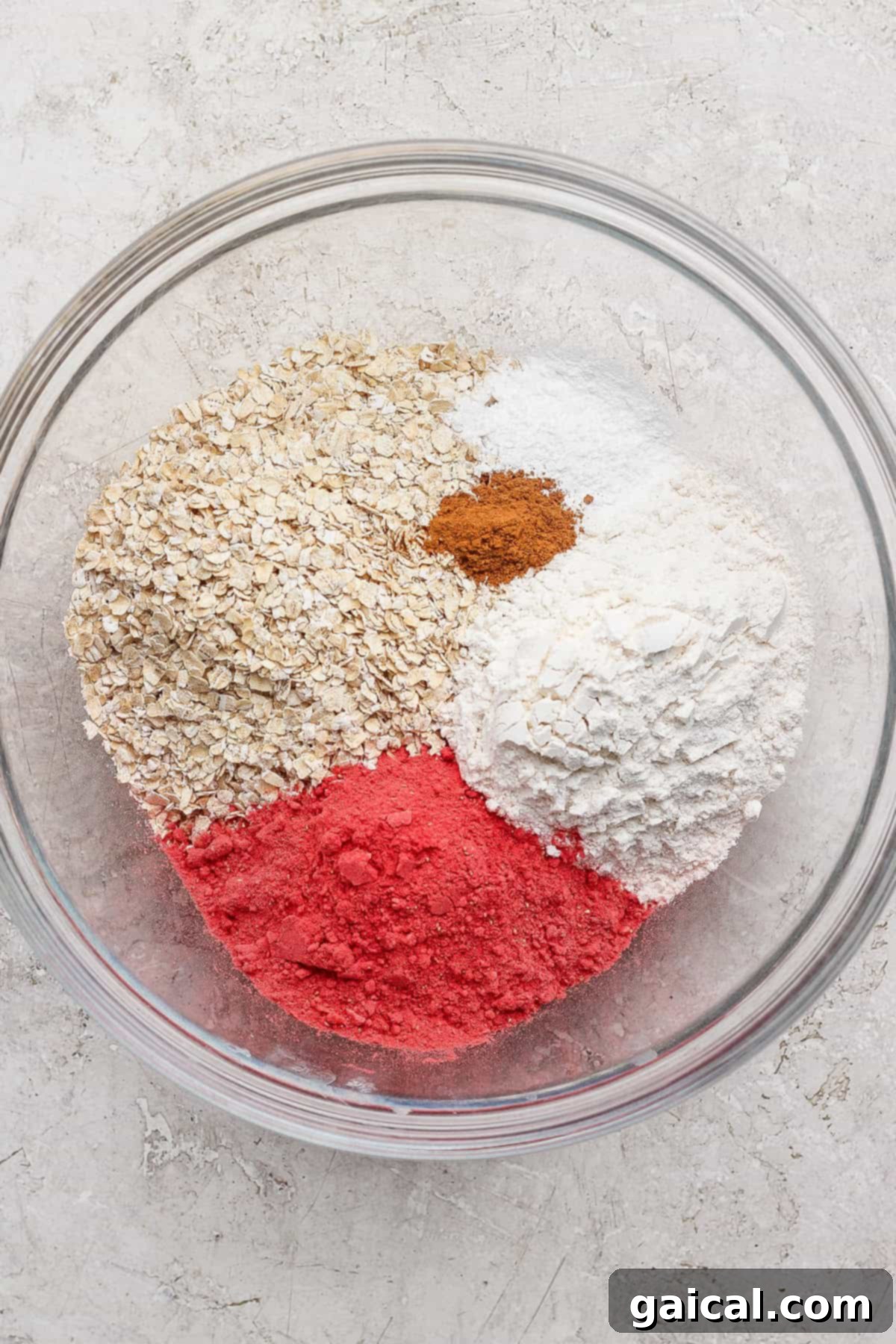 Creamy Strawberry Cheesecake Baked Oats 3 A clear glass bowl containing a vibrant mixture of dry ingredients including quick-cooking oats, white all-purpose flour, pink freeze-dried strawberry powder, cinnamon, baking powder, and baking soda, all resting on a light, textured surface.