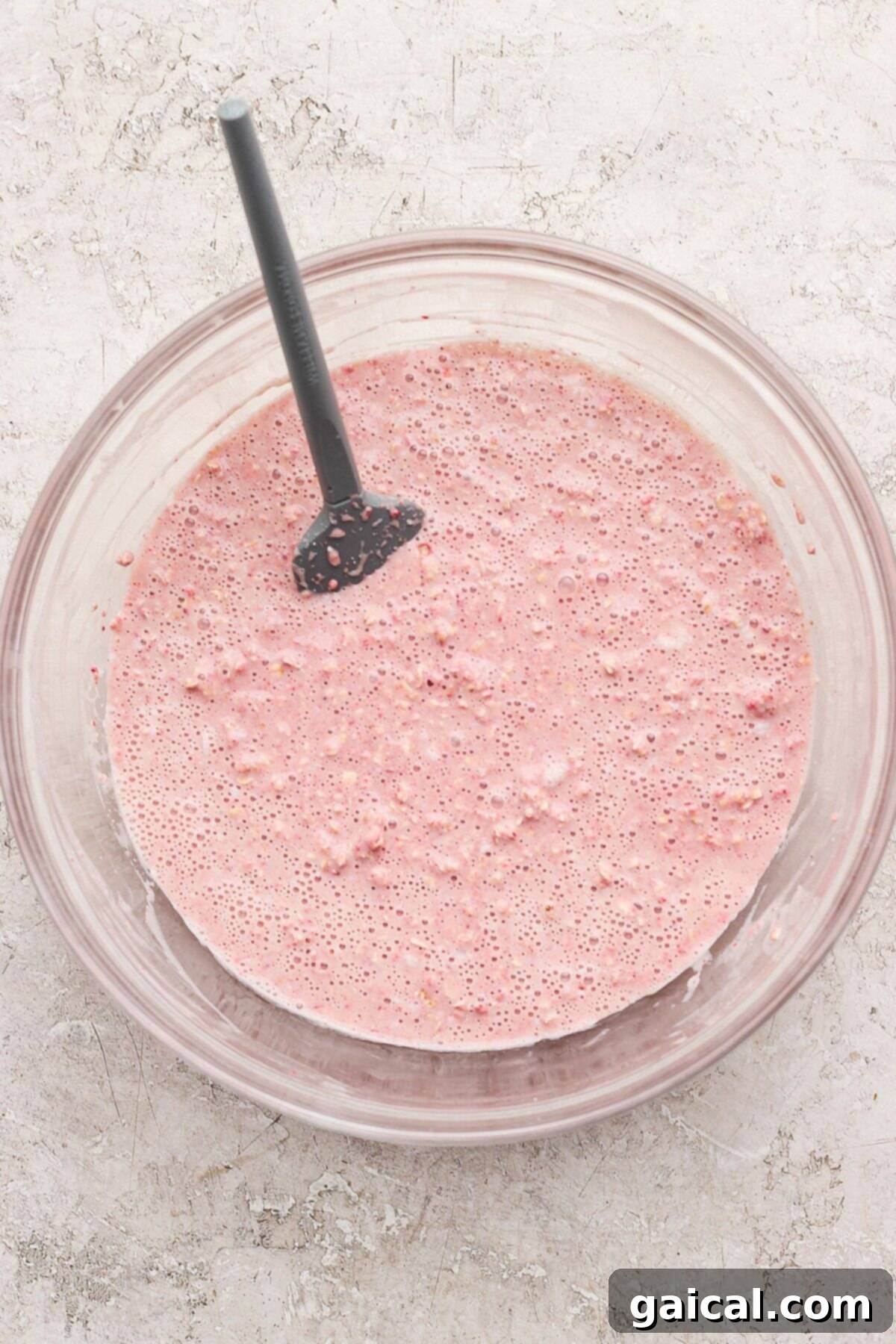 Creamy Strawberry Cheesecake Baked Oats 4 A glass mixing bowl filled with a smooth, light pink batter mixture for baked oatmeal, with a black rubber spatula resting in it, set on a light textured surface, ready for baking.