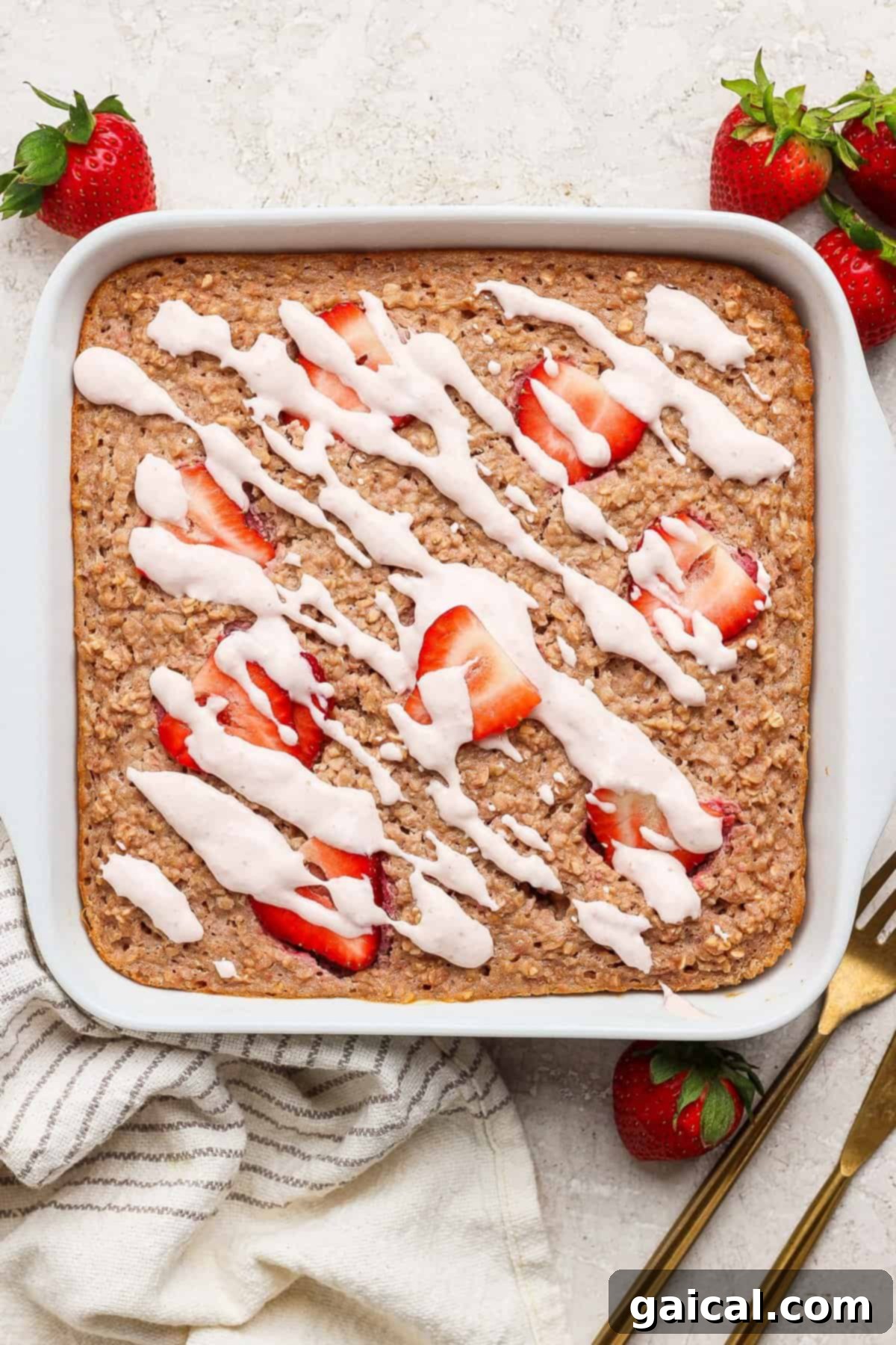 Creamy Strawberry Cheesecake Baked Oats 5 A square pan of baked oatmeal topped generously with vibrant sliced strawberries and a delicate white icing drizzle, set on a bright, light-colored surface. Nearby, whole fresh strawberries, a stylish striped towel, and elegant gold utensils complete the inviting scene, highlighting the dish's fresh appeal.