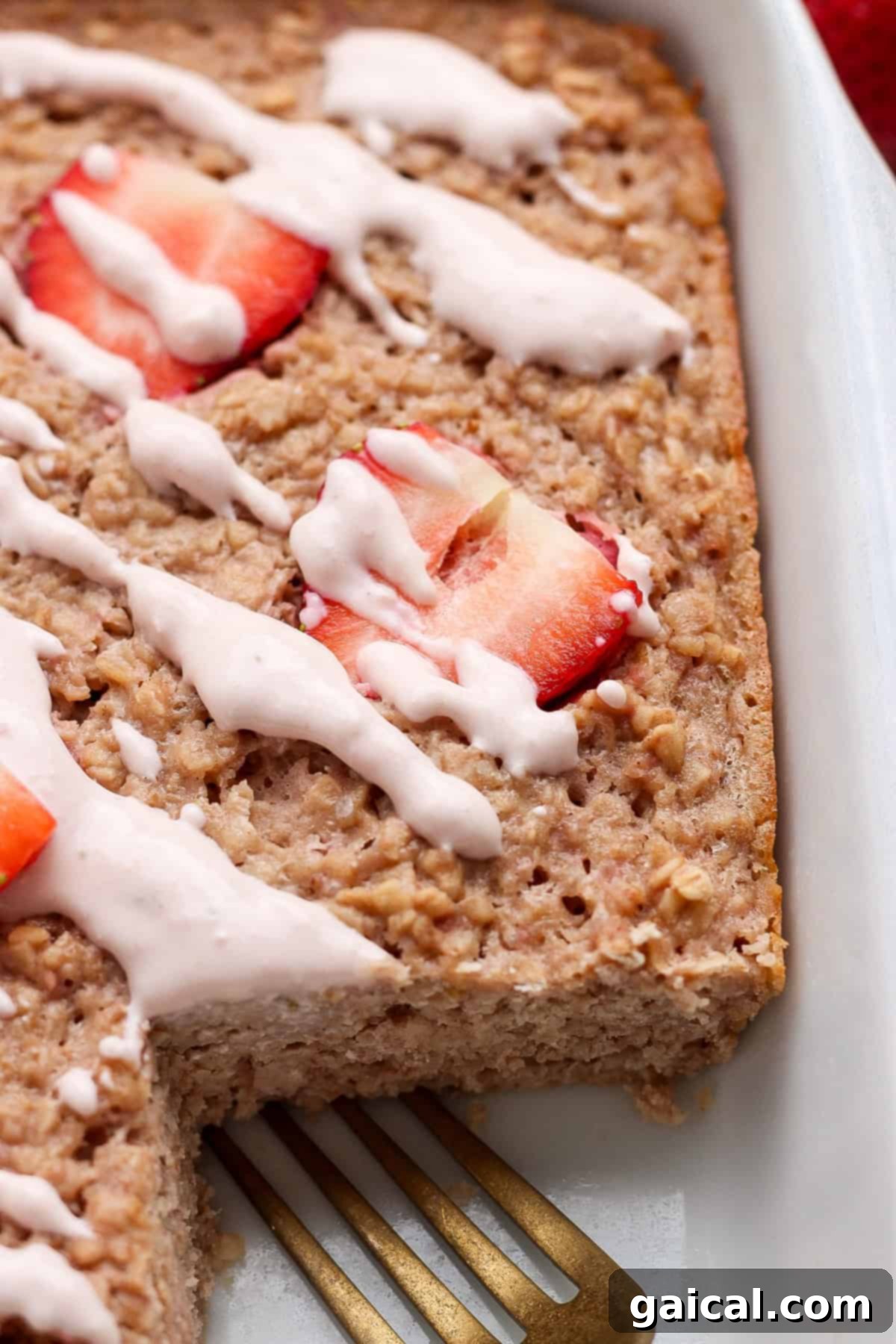 Creamy Strawberry Cheesecake Baked Oats 6 A baking dish filled with a golden-brown baked oatmeal, adorned with vibrant sliced fresh strawberries and a creamy white drizzle. A single serving has been carefully removed, showcasing the moist interior, with a sleek metal spatula resting nearby, inviting another portion.