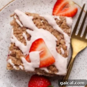 A square piece of baked oatmeal cake topped with pink icing and sliced fresh strawberries, perfectly served on a speckled plate with a gold fork, enticing a delicious bite.