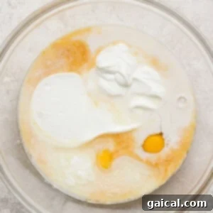A glass mixing bowl containing an egg, milk, yogurt, and other liquid baking ingredients on a light textured surface, perfectly combined to form the wet base of the recipe.