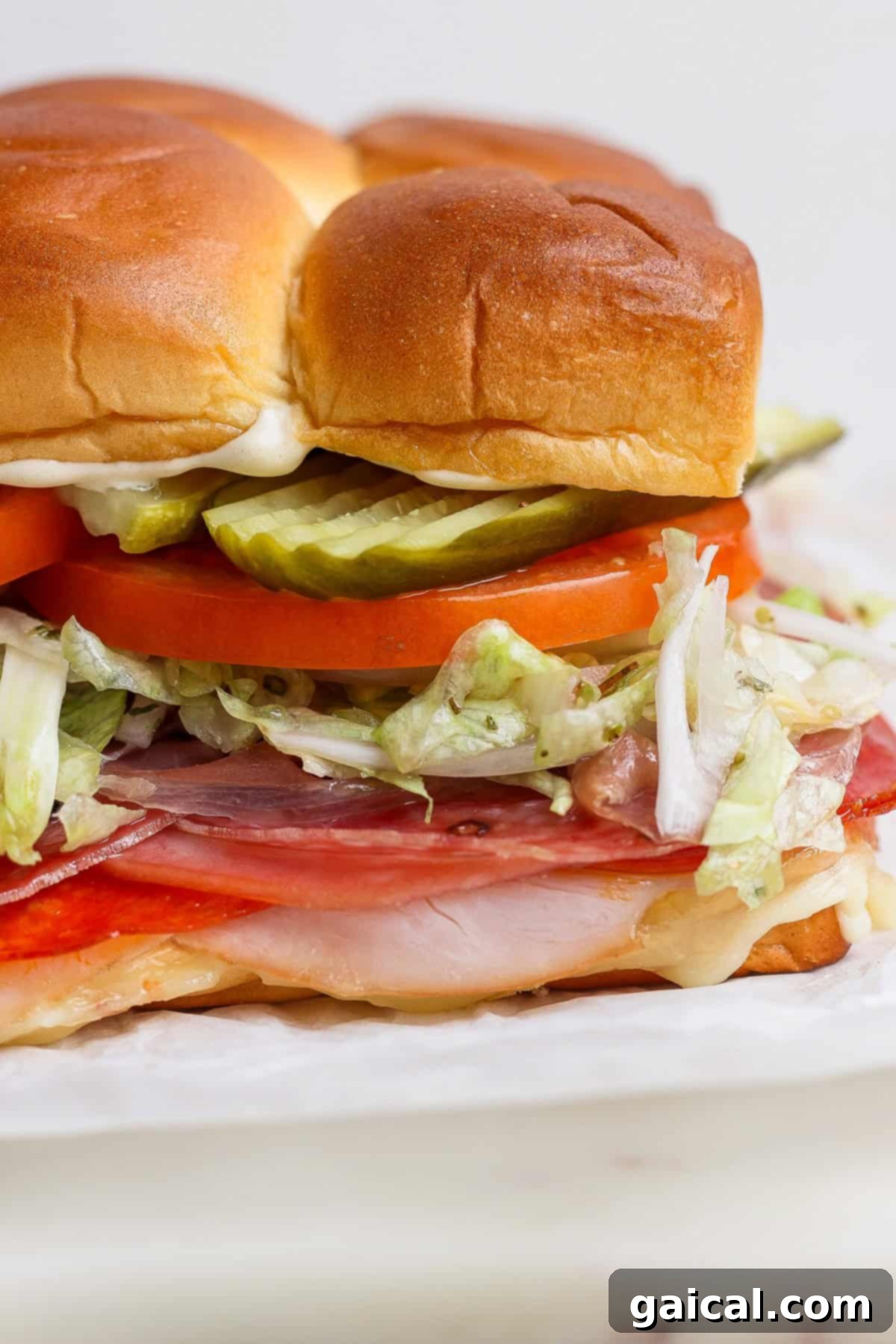 A vibrant close-up of a delectable Italian grinder slider, showcasing layers of premium sliced meats, crisp shredded lettuce, ripe tomato, tangy pickles, and a creamy mayonnaise spread, all nestled within a soft, perfectly browned Hawaiian roll. The textures and colors invite you to take a bite.
