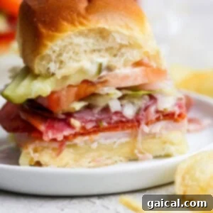 A small, mouth-watering Italian grinder slider with layers of fresh deli meats, melted cheese, crisp lettuce, juicy tomato, and pickles on a soft bun, with a bite taken out to reveal its delicious fillings. This image captures the essence of a perfect, easy-to-eat party snack, served with potato chips.