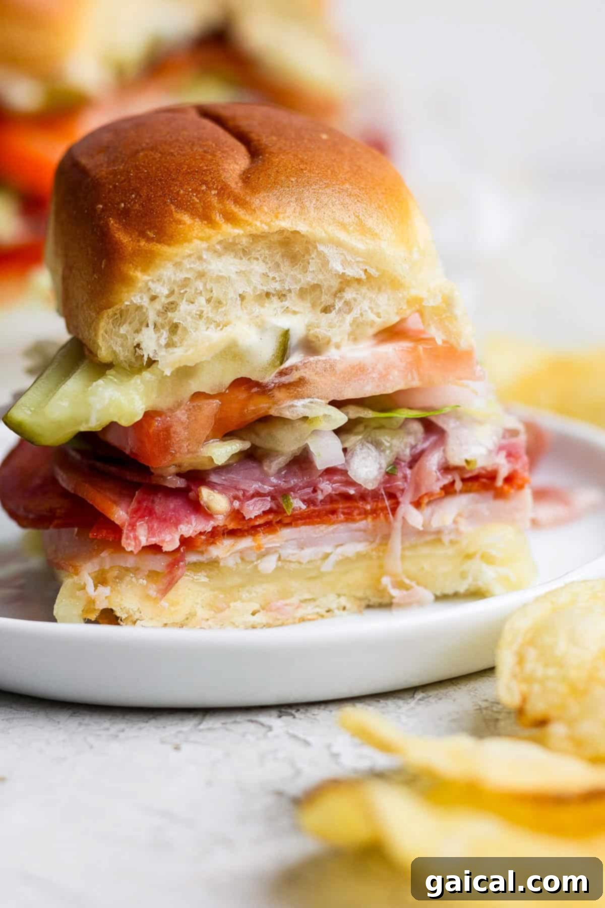 A single, perfectly cut Italian Grinder Slider presented on a pristine white plate, showcasing its rich layers of savory deli meats, gooey melted cheese, fresh shredded lettuce, juicy tomato slices, and crisp pickles. A small heap of golden, crunchy potato chips accompanies the slider, with a corner visibly taken from the sandwich, inviting the viewer to indulge.