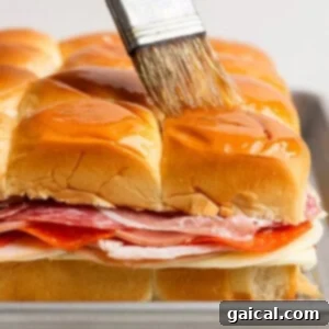 A hand brushes melted butter over the tops of stacked sandwich sliders with layers of deli meats and cheese on a baking tray. This step ensures a golden, crispy crust.