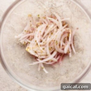 A clear glass bowl containing thinly sliced red onions mixed with dried herbs, specifically oregano, and red wine vinegar on a light, textured surface. This is the first step in preparing the signature zesty topping.