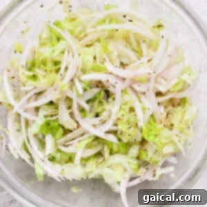 A clear glass bowl showcasing shredded lettuce and thinly sliced onions, thoroughly mixed with olive oil, black pepper, and seasonings, sitting on a light textured surface. This mixture forms the fresh, zesty topping for the Italian grinder sliders.