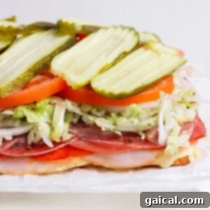 The baked Italian Grinder Sliders on a cutting board, with the top layer of Hawaiian rolls gently removed to reveal the melted cheese and deli meats. The seasoned lettuce, sliced tomatoes, and pickles are being added as fresh toppings, preparing the sliders for the final step.