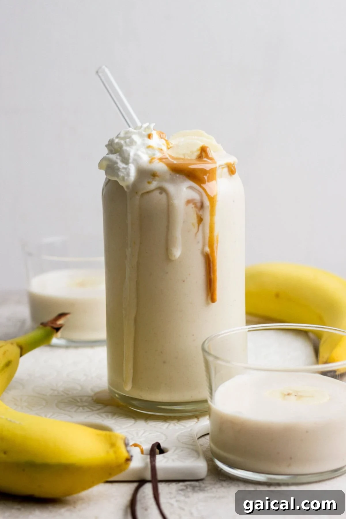 Glass of banana smoothie topped with whipped cream and peanut butter, surrounded by bananas and two small glasses of smoothie.