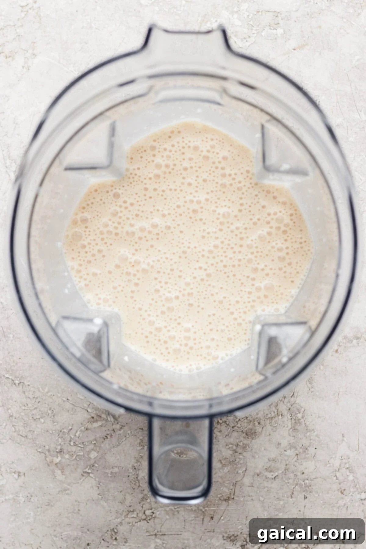 Top-down view of a blender containing a light beige, frothy liquid on a light textured surface.