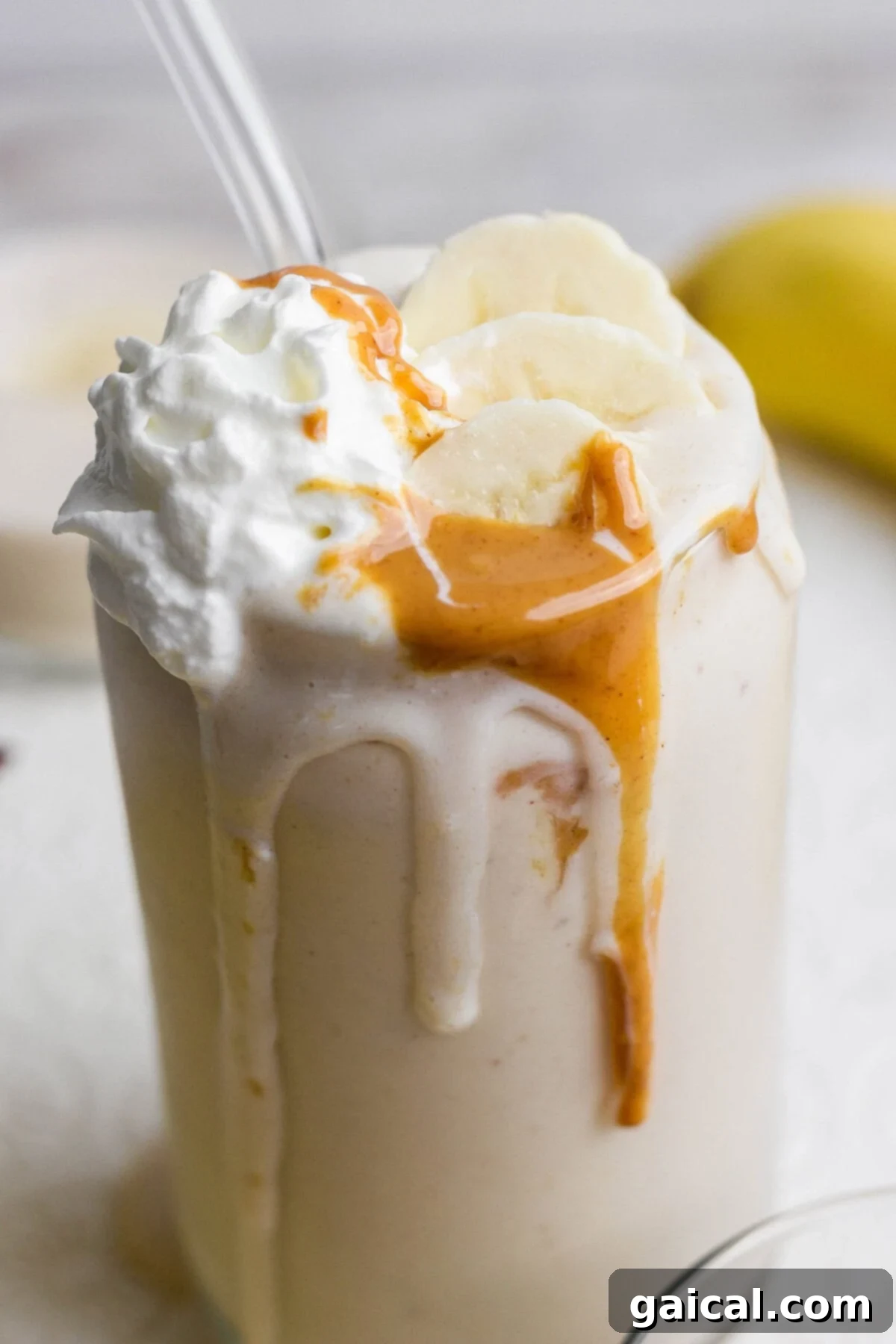 A glass of banana milkshake topped with whipped cream, banana slices, and peanut butter drizzle, with a straw inserted.