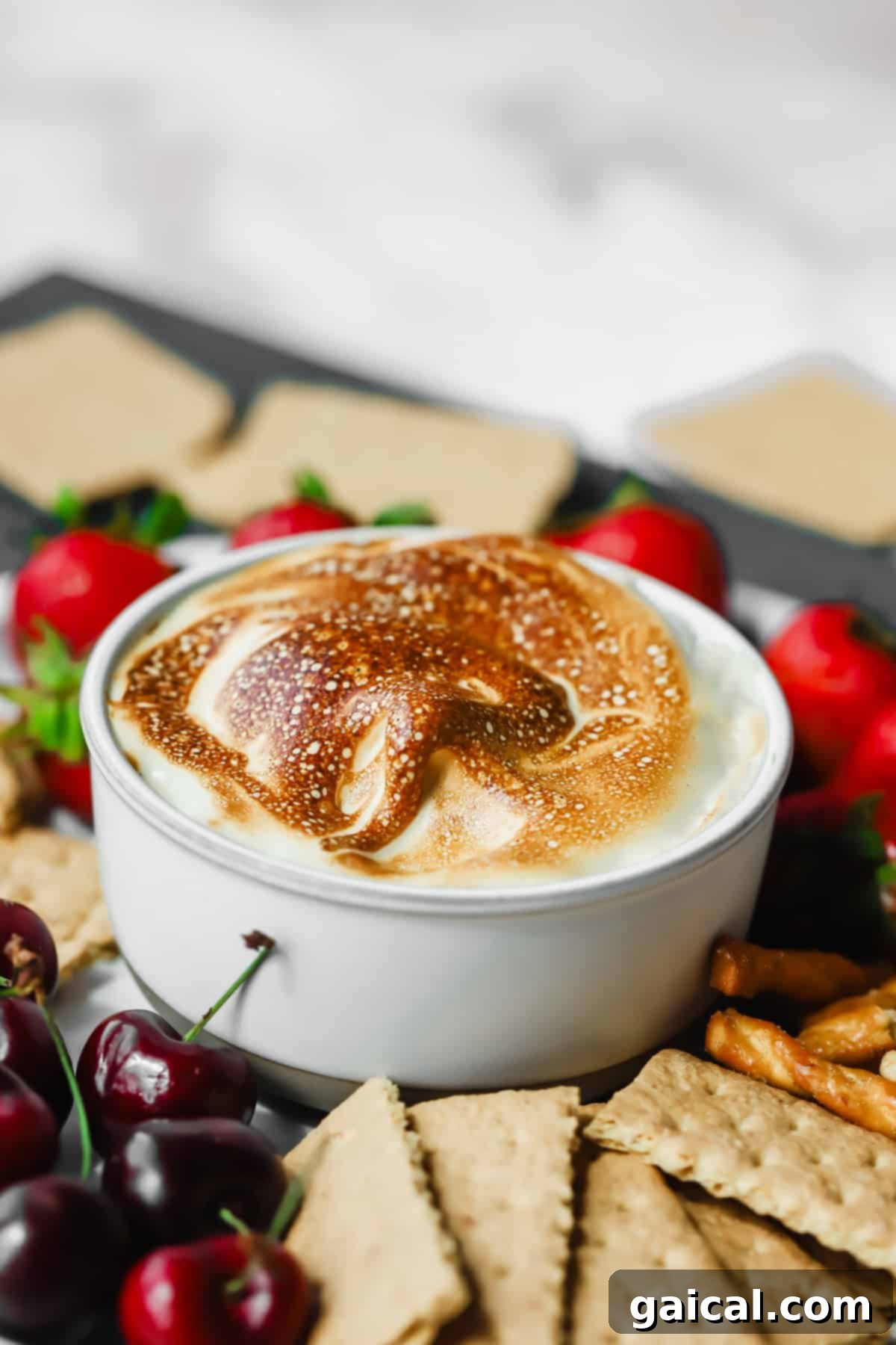 Maple Mallow Cloud 2 A white bowl filled with toasted marshmallow dip, surrounded by graham crackers, pretzels, strawberries, and cherries.