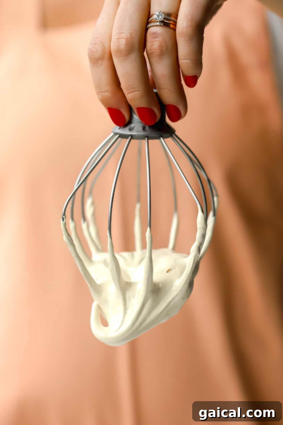 Maple Mallow Cloud 4 A hand with red nail polish holds a metal whisk coated in thick, creamy batter against a peach-colored background.