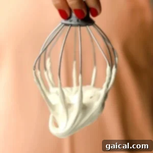 A hand with red nail polish holds a metal whisk covered in thick, maple marshmallow fluff against a peach-colored background.