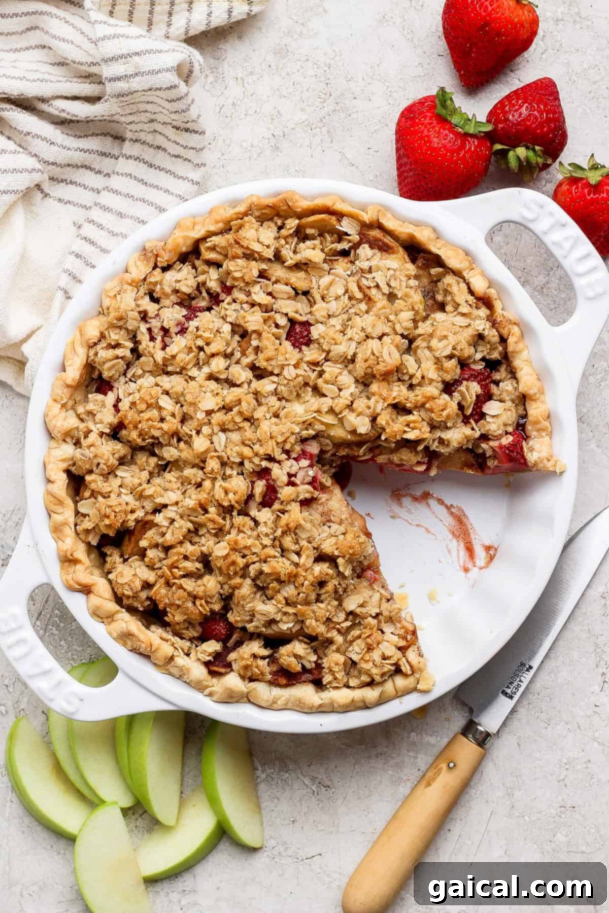 Sun-Kissed Strawberry Apple Pie 3 A vibrant strawberry apple pie, adorned with a golden oat crumble topping, nestled in a pristine white pie dish. One inviting slice has been removed, revealing layers of luscious fruit. Fresh strawberries, crisp apple slices, and a gleaming knife are artfully arranged nearby.