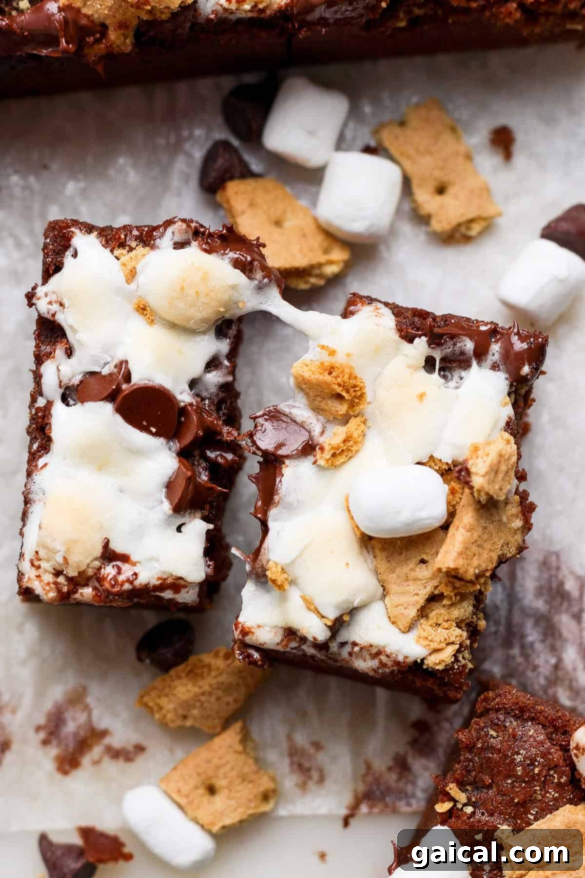 Two s'mores brownies topped with melted marshmallows, chocolate chips, and graham cracker pieces, placed on parchment paper with scattered ingredients around.